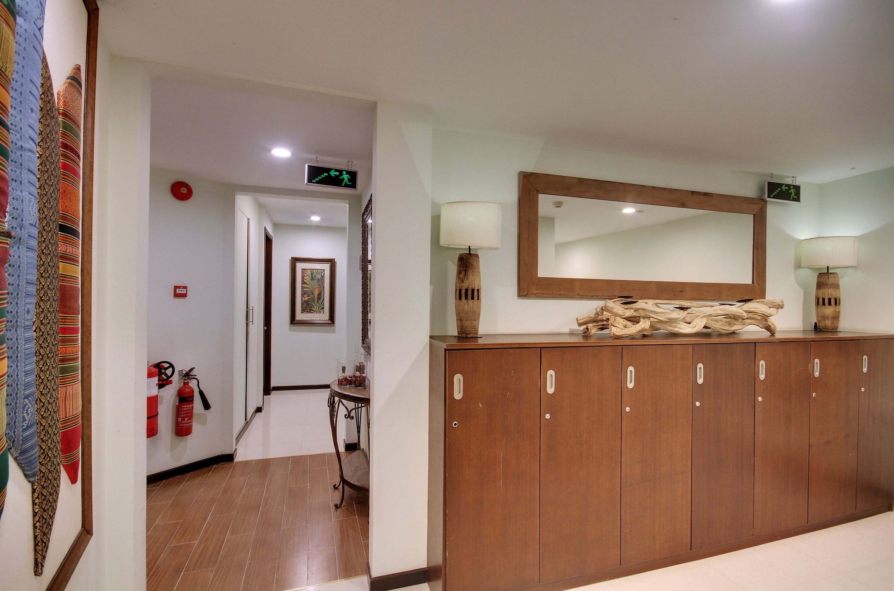A hallway with a wooden wardrobe, a mirror, and a table with a vase. There is a fire extinguisher on the wall and a door with an exit sign.