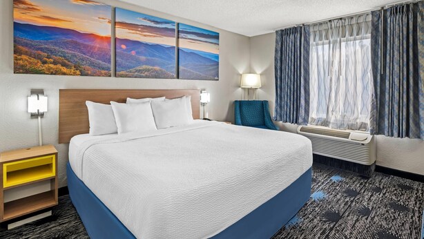 Una habitació d'hotel amb un llit gran, dues làmpades subjectes a la paret, una tauleta de nit, una finestra amb cortines i una paret amb tres quadres de paisatges.