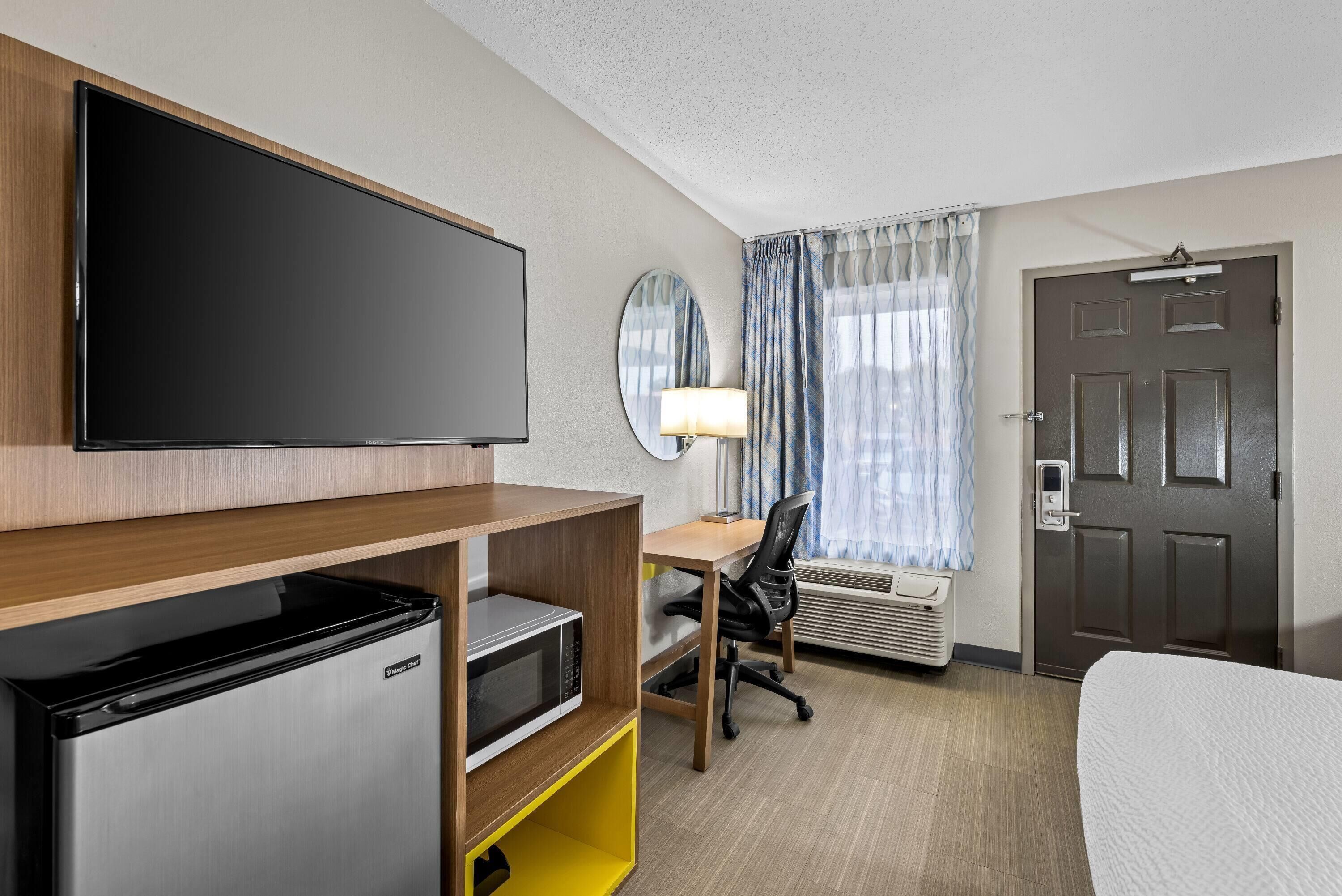 Habitación de hotel con televisión de pantalla plana, microondas, escritorio con silla, cama, ventana con cortinas y puerta con picaporte.