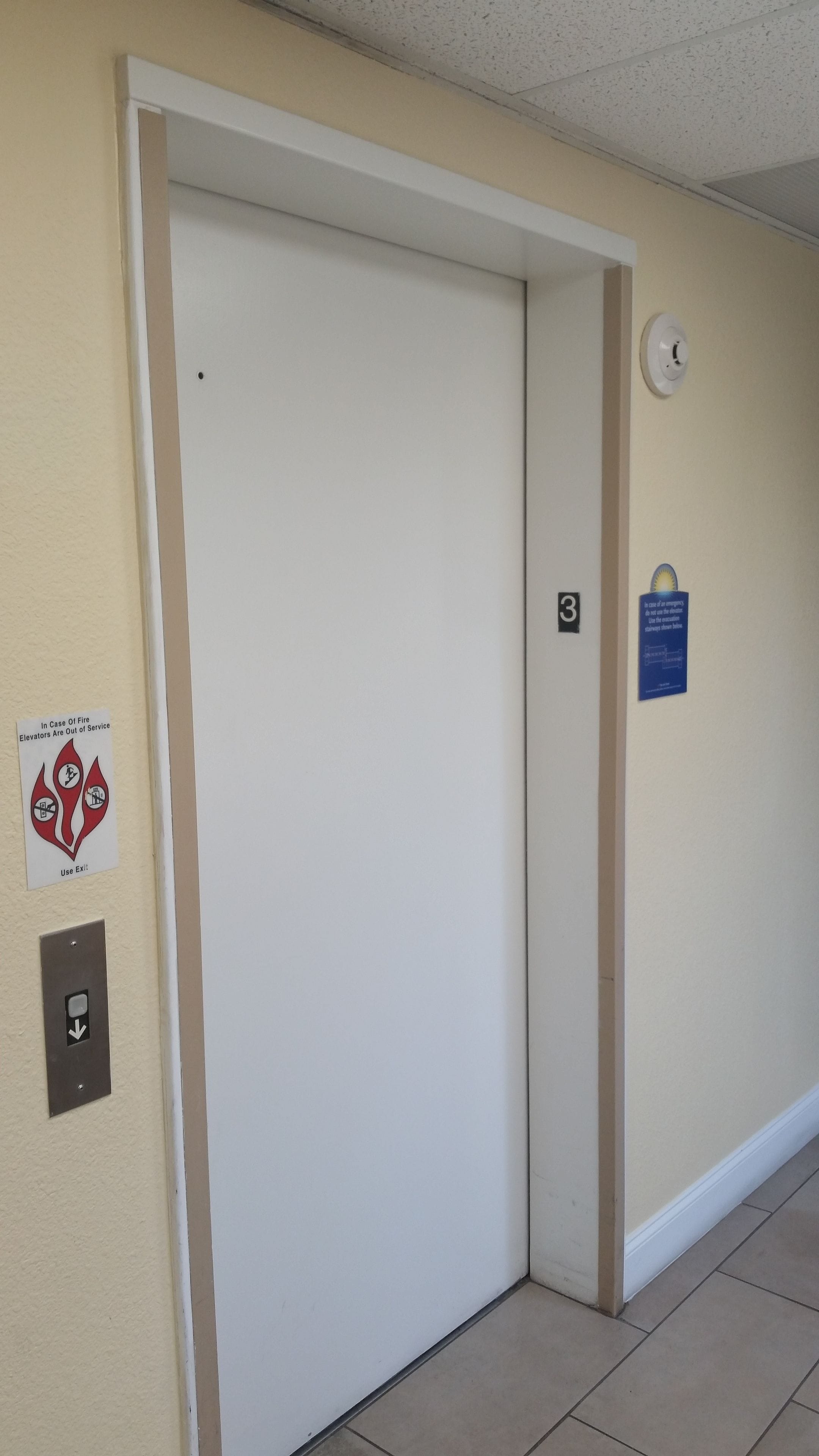 Un pasillo con una puerta de ascensor cerrada, un letrero de seguridad contra incendios y un detector de humo en la pared.