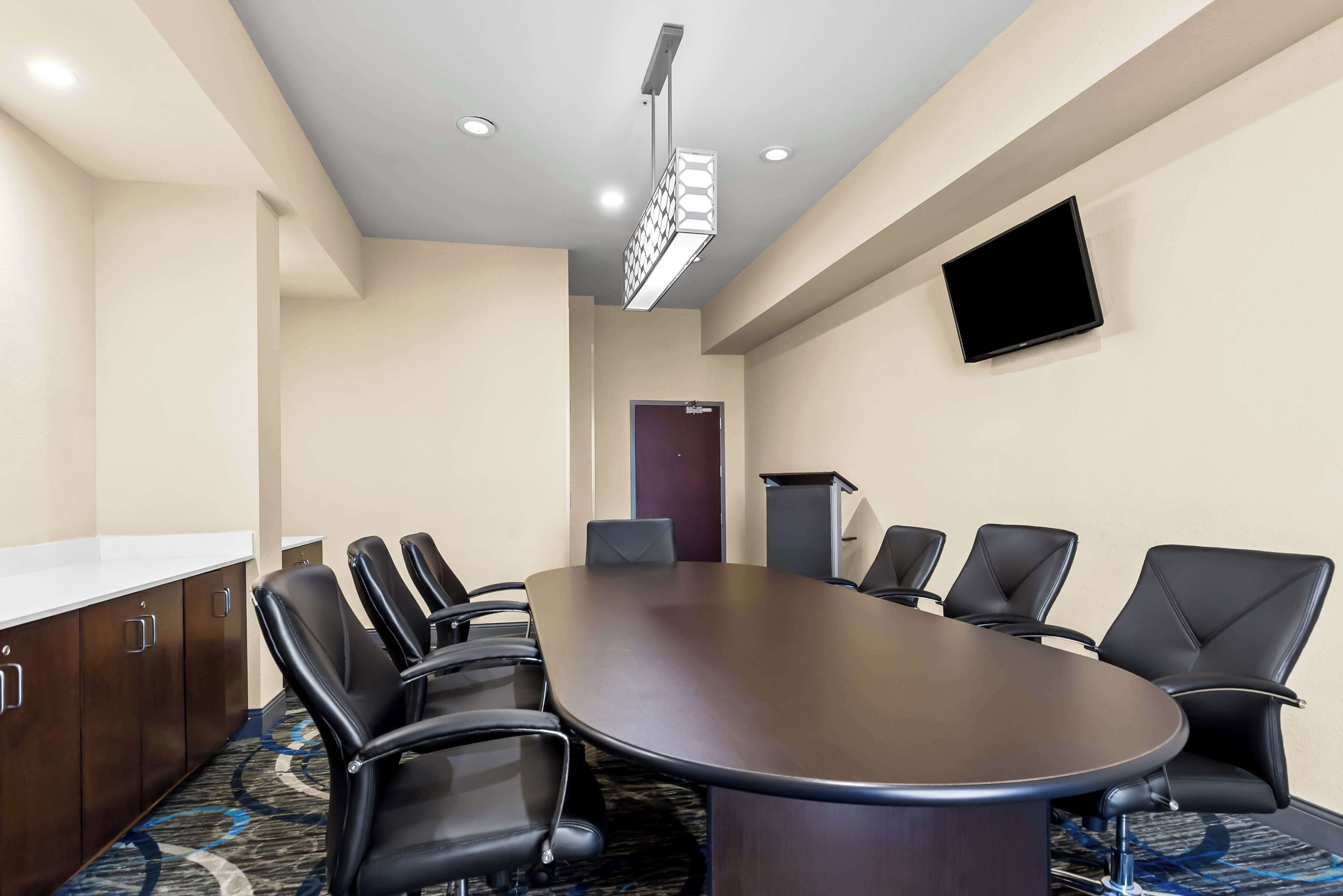 A conference room with a large oval table, black chairs, a podium, and a flat-screen TV on the wall.