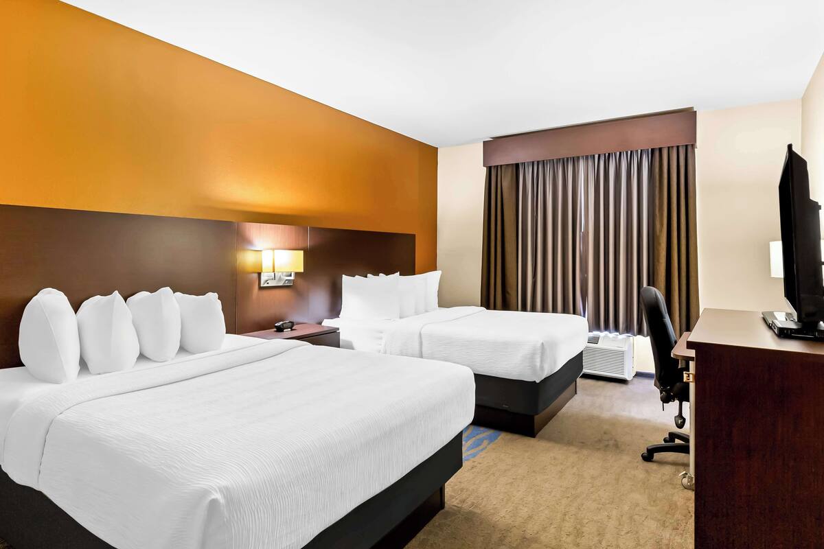 A hotel room with two beds, a desk, a chair, a lamp, and a large window with curtains.