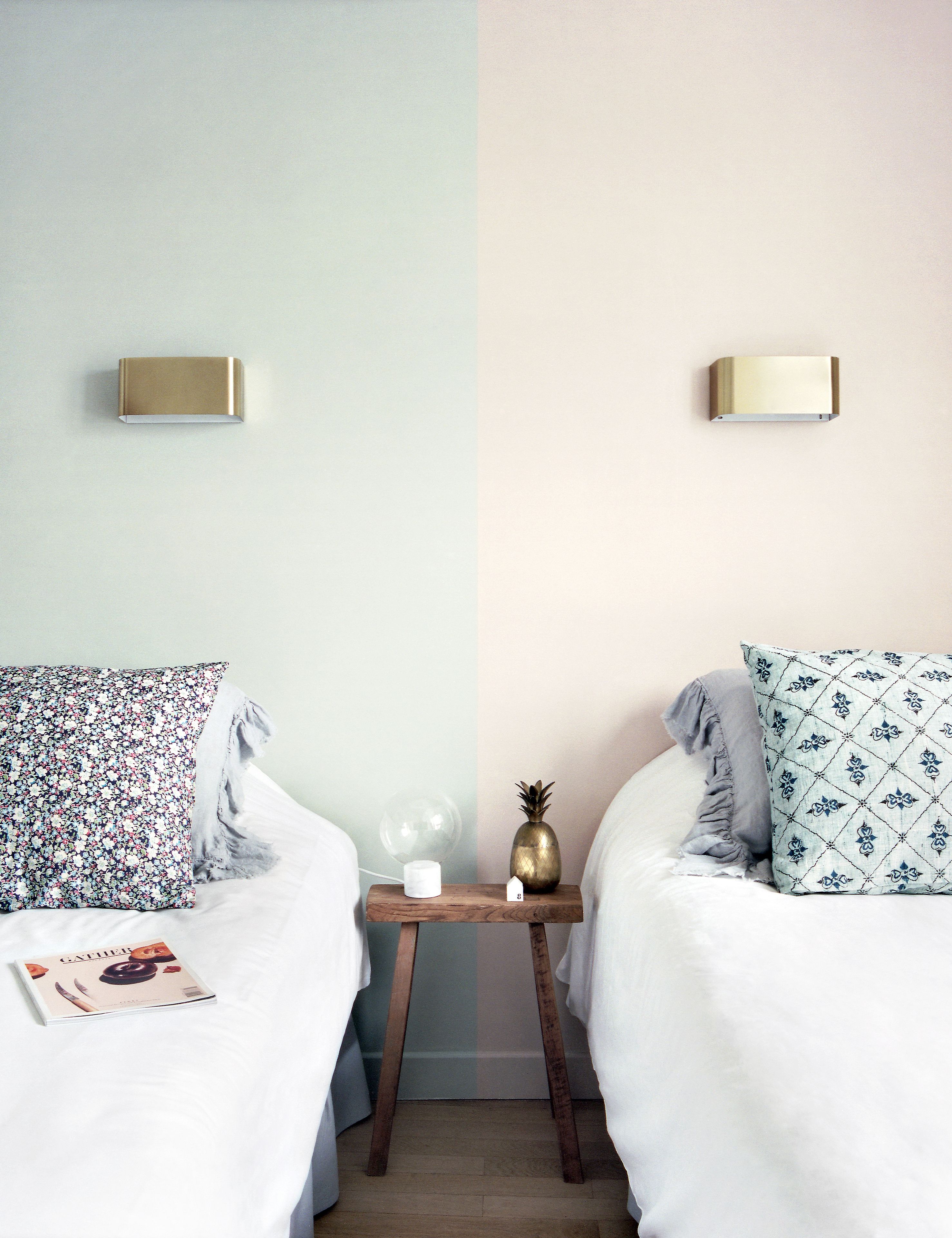Une vue partagée d’une chambre à coucher avec deux lits, chacun orné d’oreillers à motifs différents, une table de chevet avec un vase et un petit objet en forme de maison, et des appliques murales.