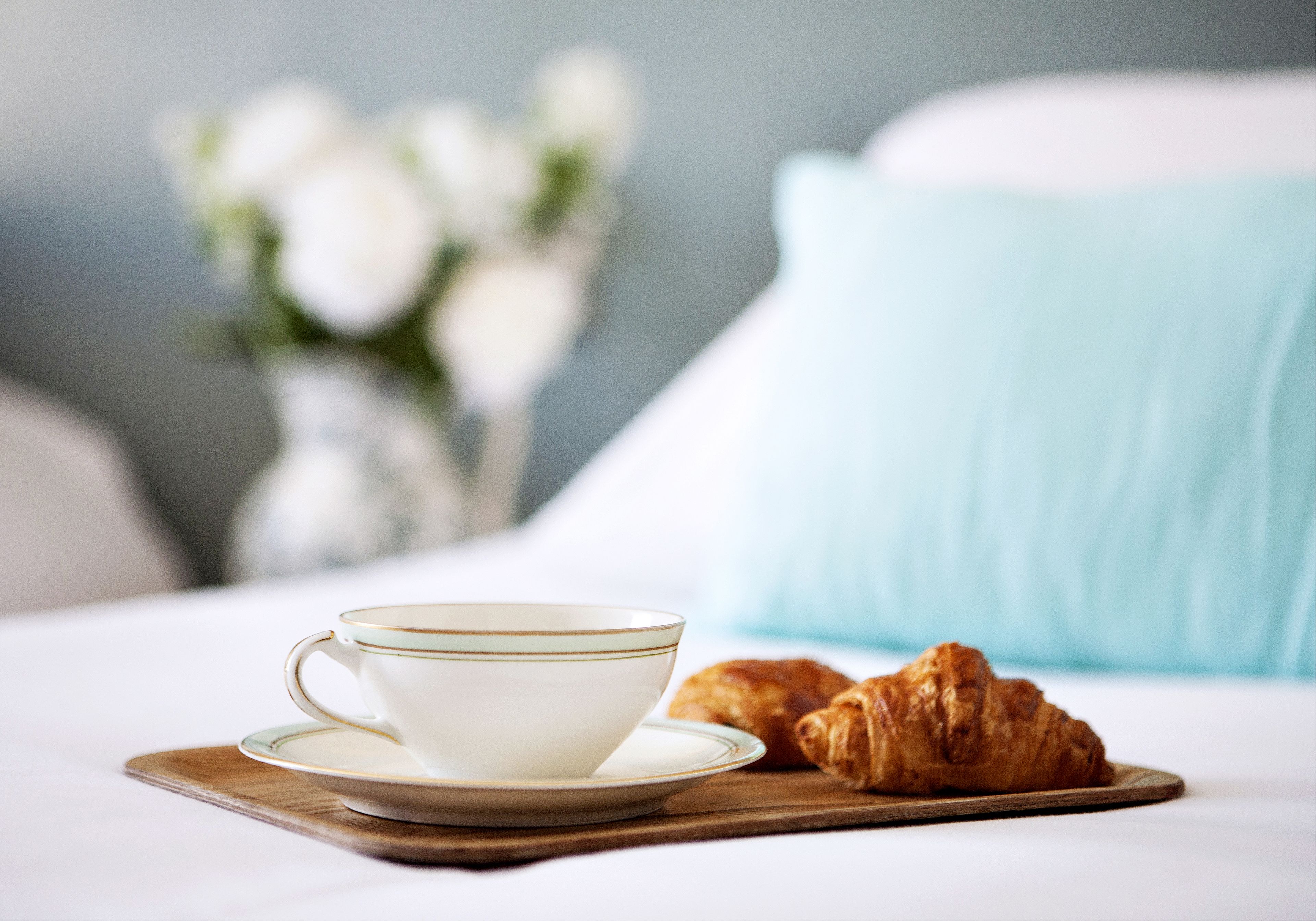 Una taza de café y croissants sobre una bandeja de madera en una cama con un cojín azul claro.
