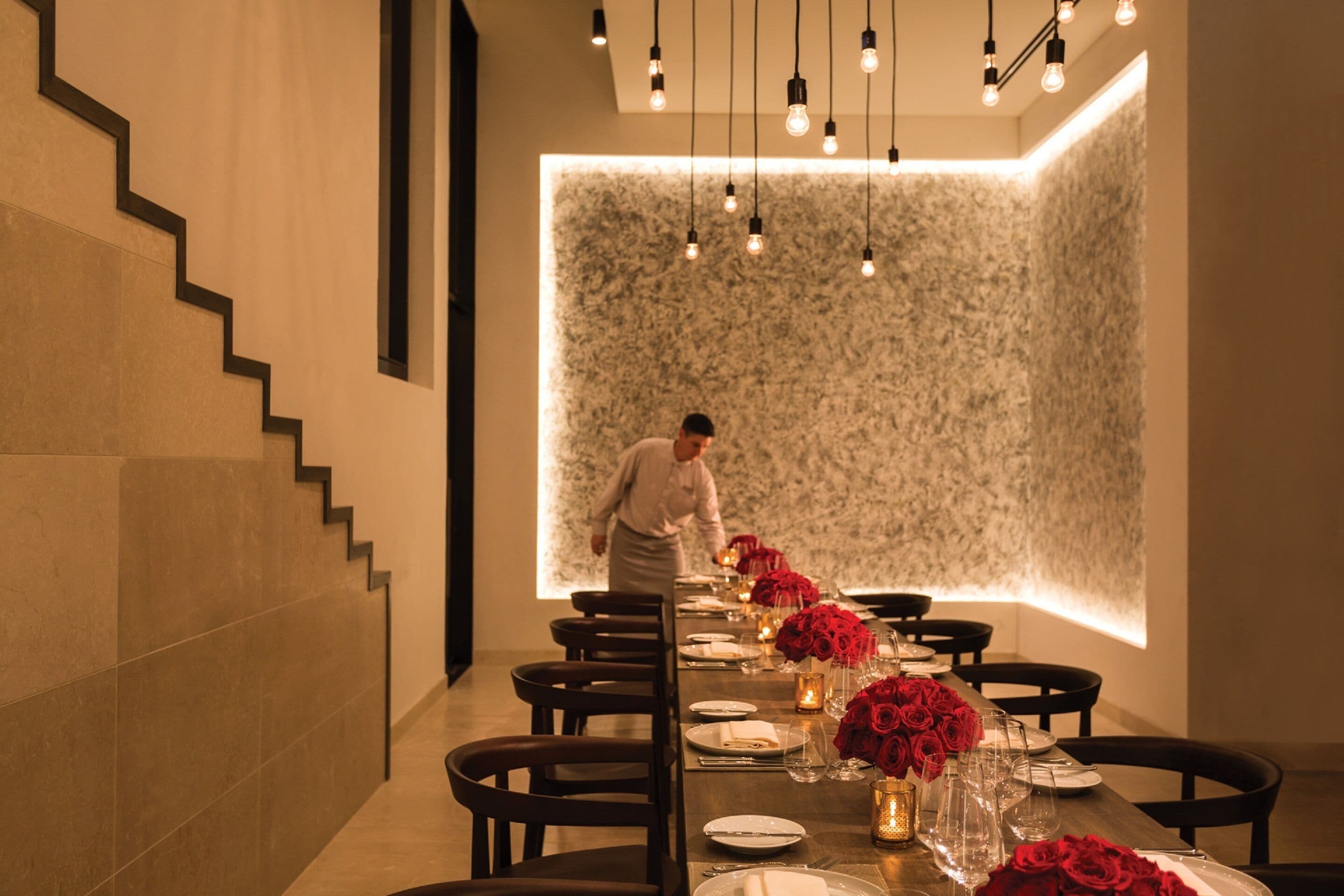 A dining area with a long table set for a meal, a staircase on the left, and a textured wall in the background.