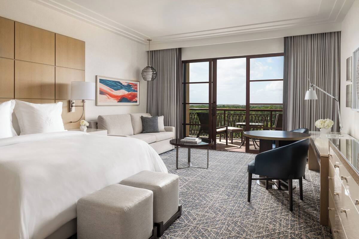 Habitación de hotel con una cama grande, un sofá, una mesa de comedor y un balcón con vistas.