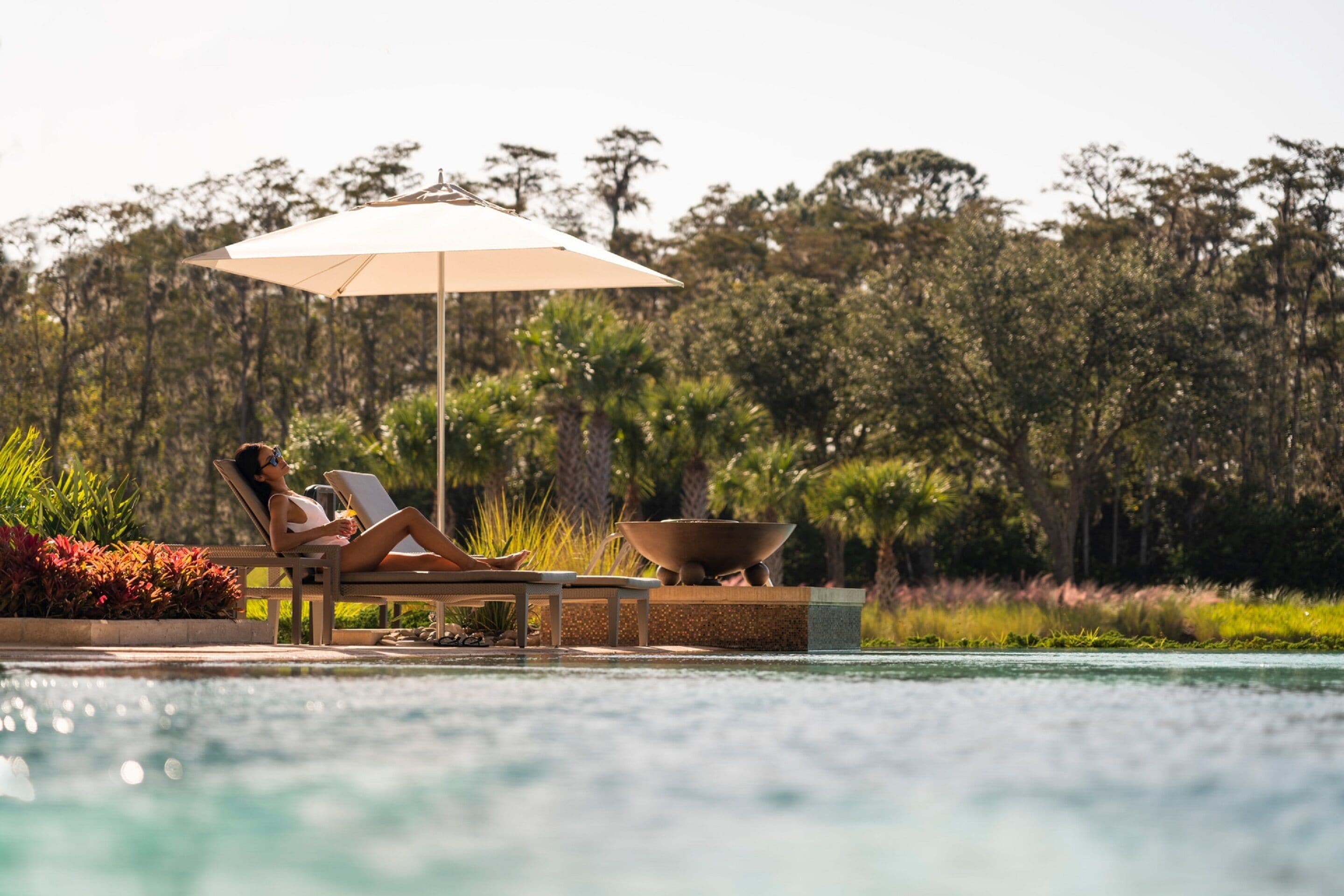 A person relaxing on a sun lounger by a pool with a large umbrella, surrounded by lush greenery.
