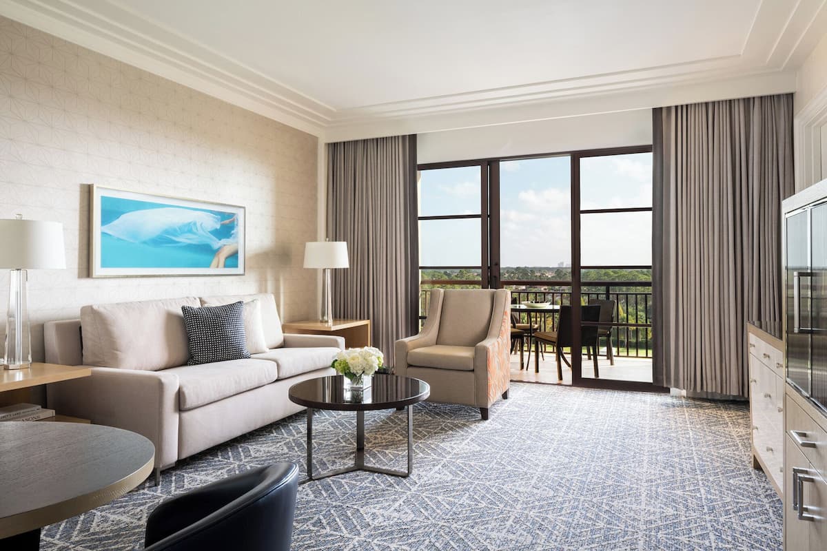 Habitación de hotel con sofá, sillón, mesa de comedor y balcón con vistas.