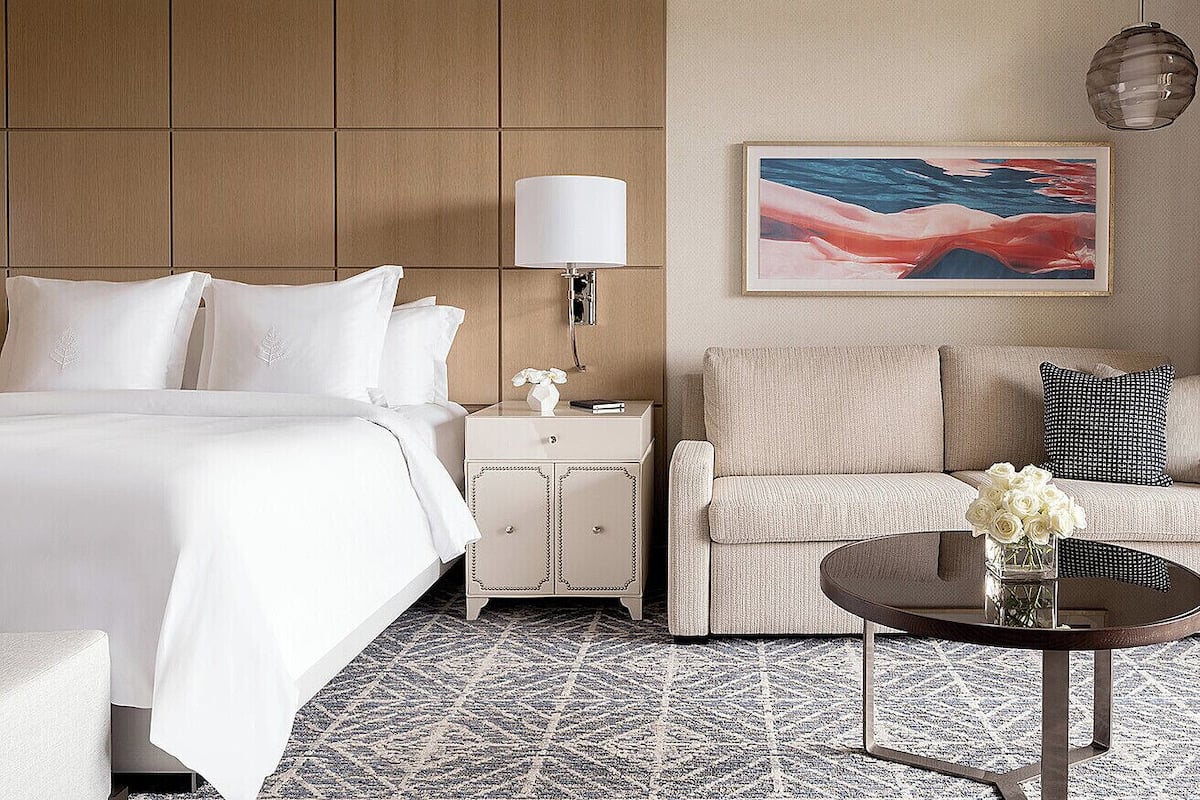 Una habitación de hotel con una cama grande, una mesita de noche con lámpara, un sofá, una mesa de centro con flores y un cuadro en la pared.