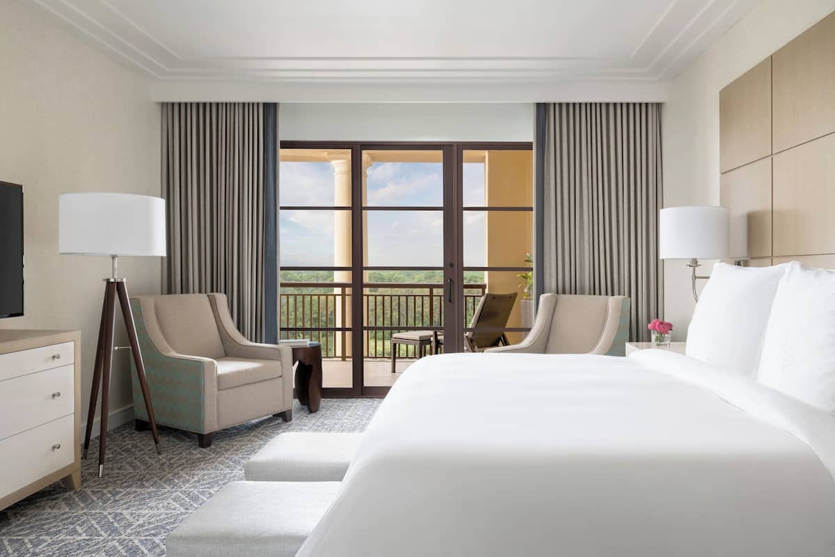 Habitación de hotel con una cama grande, dos sillones, una mesita y un balcón con cortinas.