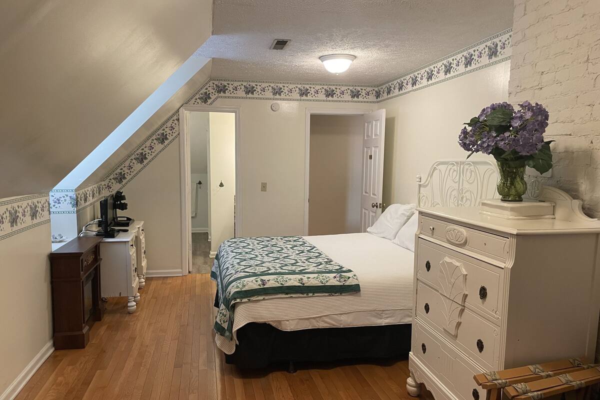 A bedroom with a bed, dresser, and a desk with a computer. There is a door leading to another room and a window with a view of the outside.