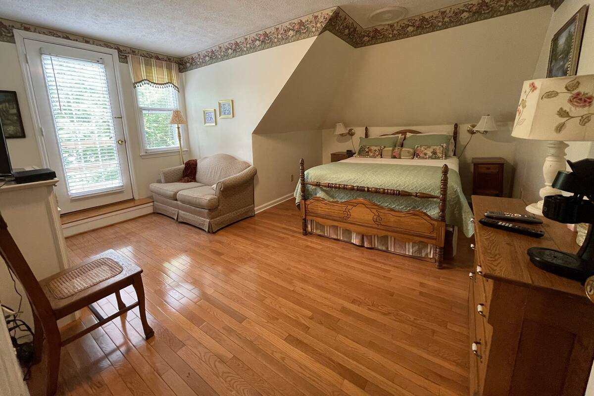 A bedroom with a wooden floor, a bed with a green bedspread, a chair, a desk with a lamp, and a window with blinds.