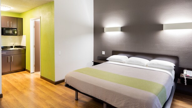 A hotel room with a bed, a nightstand, a wall-mounted light, and a kitchenette with a microwave. - Studio 6 Cypress, CA (Cypress)