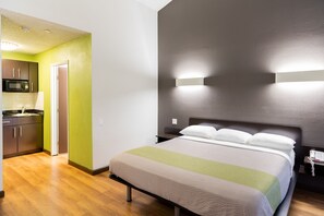 A hotel room with a bed, a nightstand, a wall-mounted light, and a kitchenette with a microwave. - Studio 6 Cypress, CA (Cypress)