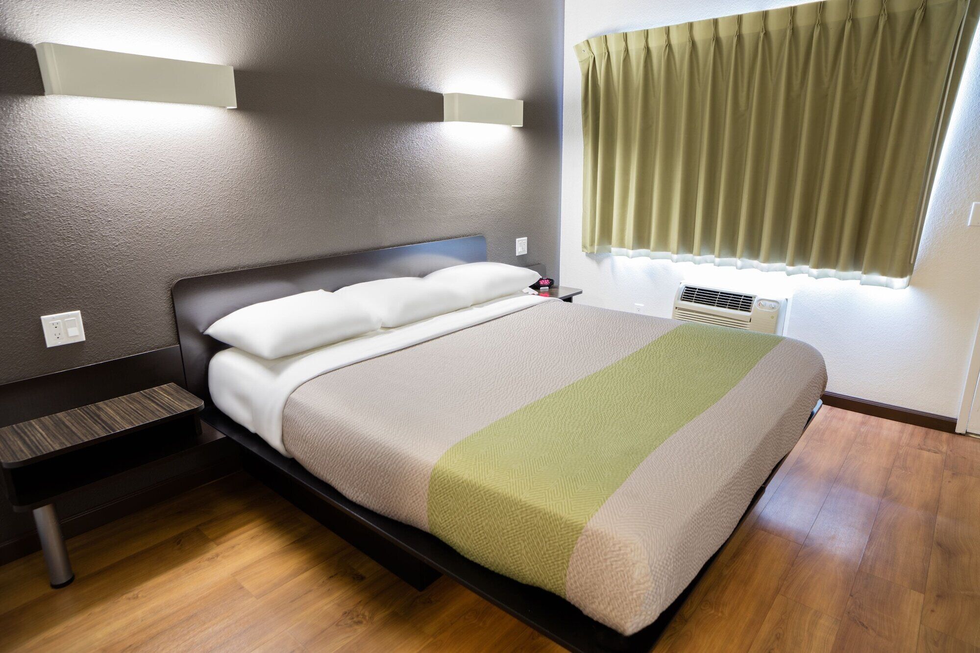 A neatly made bed with a headboard, a bedside table, a power outlet, and a window with curtains.