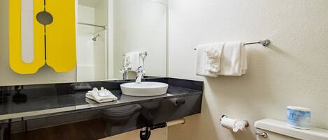 A bathroom with a black countertop, white sink, and a mirror. There is a toilet with a paper towel holder and a tissue box.