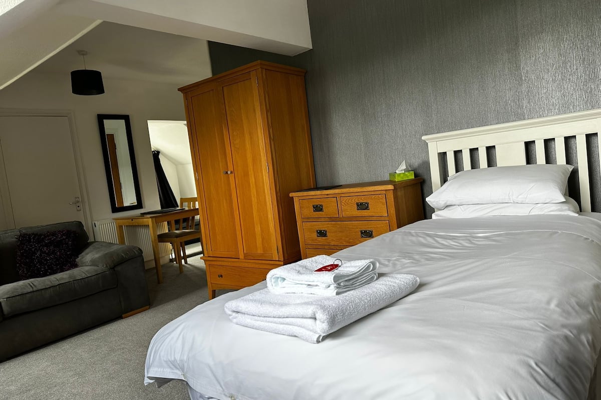 A bedroom with a bed, a wooden wardrobe, a grey sofa, and a desk with a chair.