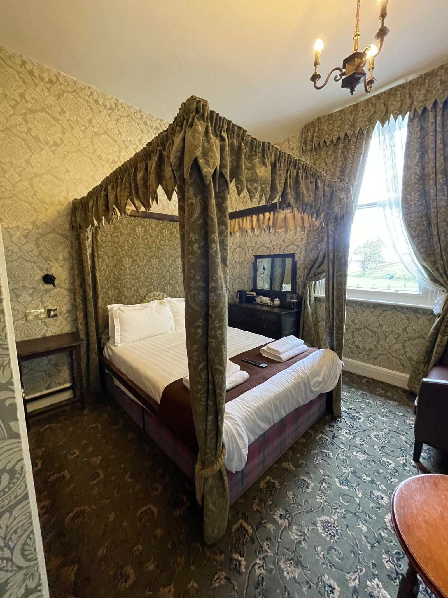 A four-poster bed with a canopy, a bedside table with a mirror, and a view of the outdoors through a window with curtains.