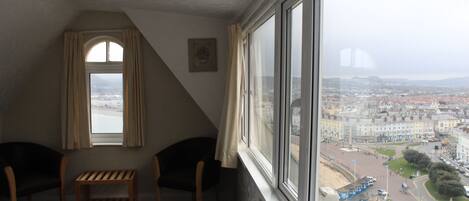 A hotel room with a bed, two chairs, a small table, and a large window offering a city view.