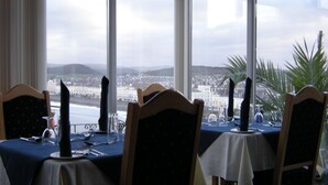 A dining area with tables set for two, overlooking a coastal cityscape through large windows. - Headlands Hotel (Llandudno)