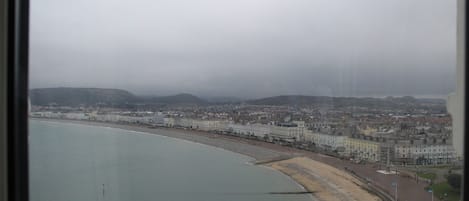 Paisagem costeira com uma praia extensa, edifícios à beira-mar e vista para colinas ao longe.