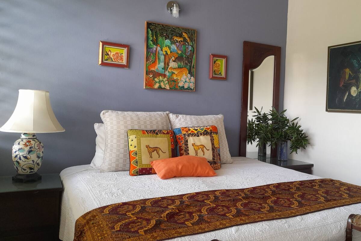 Een slaapkamer met een bed, kussens, een lamp, een spiegel en decoratie aan de muur.