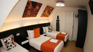 Habitación de hotel con tres camas dispuestas en fila, cada una con ropa de cama blanca y detalles naranja. Se observan pinturas abstractas en la pared sobre las camas, y un mueble negro a la derecha.