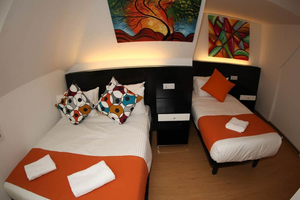 A hotel room with two beds, each with an orange bedspread and white pillows. There are colorful pillows on one bed, and a painting is visible on the wall above the beds.