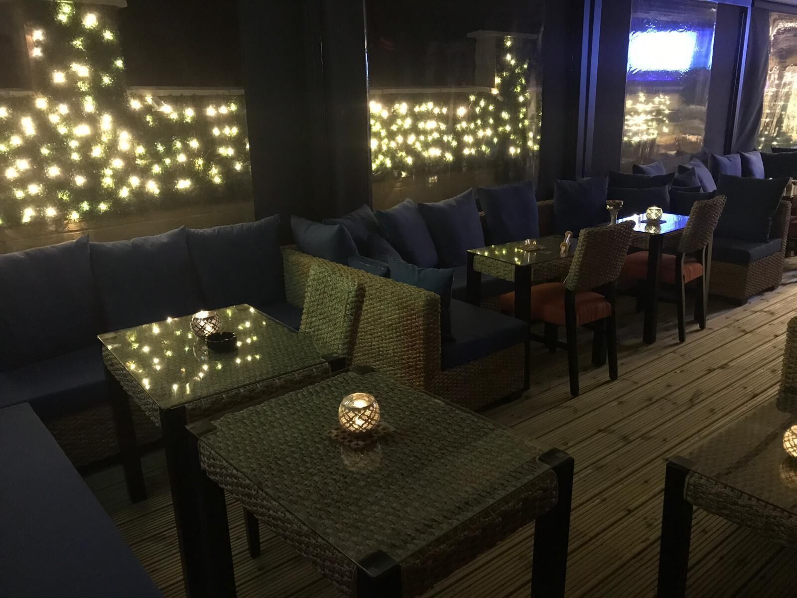 A cozy indoor seating area with cushioned sofas and tables, illuminated by string lights and candles.