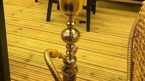 Un narguile con base de metal y una manguera, sobre un piso de madera. En el fondo, un sofá con cojines azules y una silla de mimbre.
