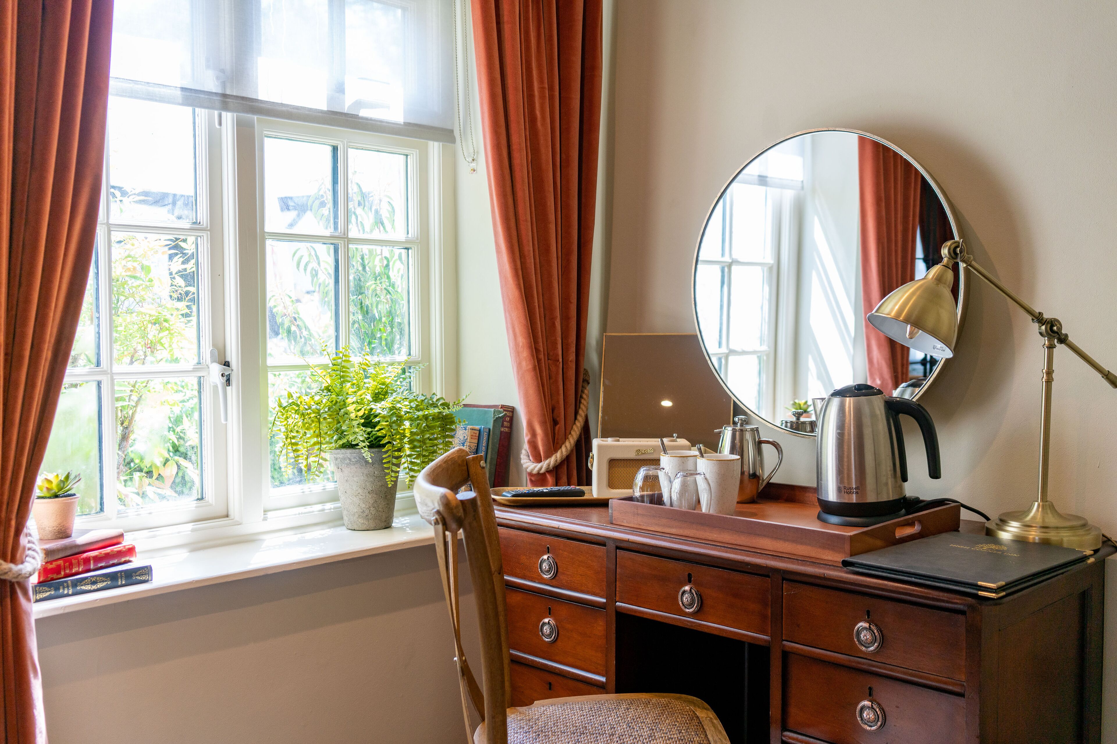 Een kamer met een houten dressoir, een spiegel, een bureaulamp, een waterkoker en een raam met gordijnen.