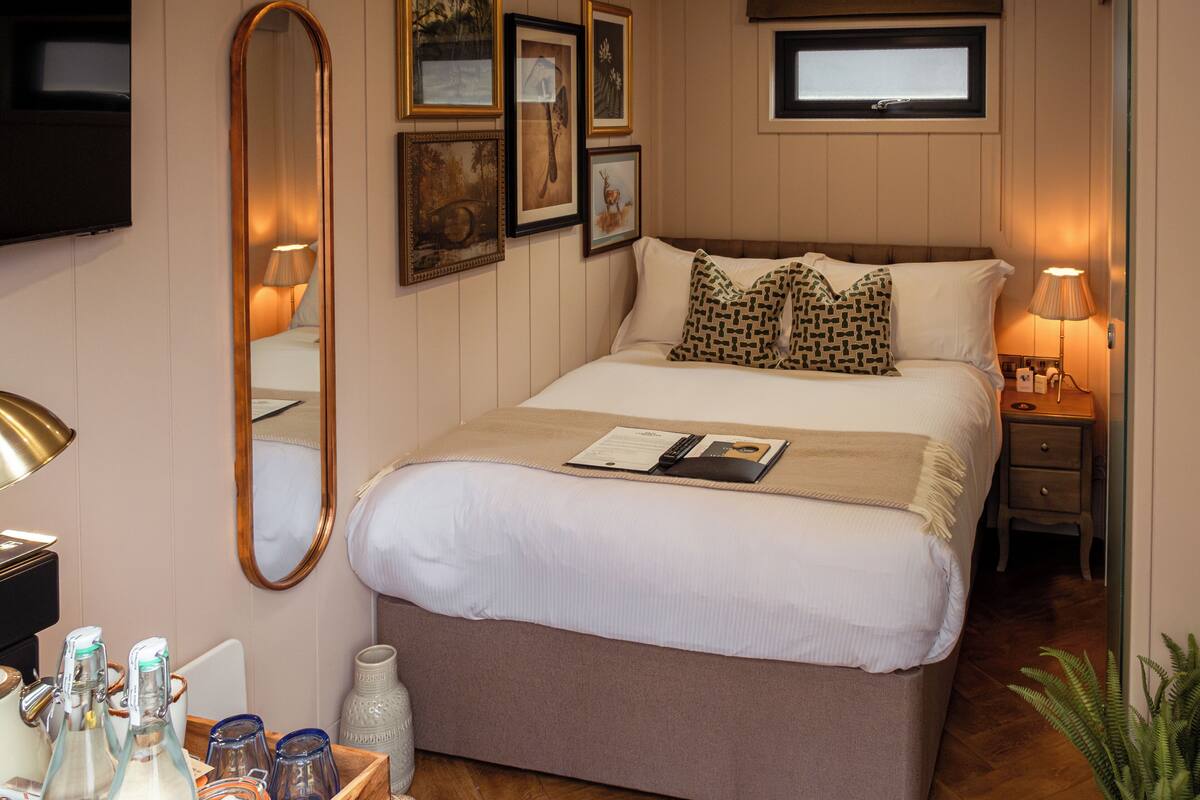 A neatly arranged hotel room with a large bed, a bedside table with a lamp, a small window, and wall-mounted artwork.