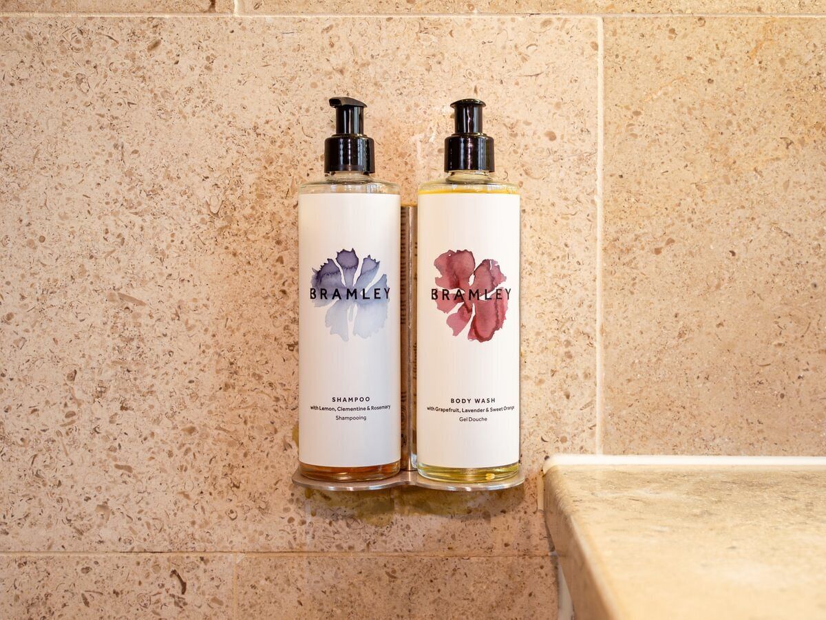 Twee flessen Bramble shampoo en douchegel op een beige betegelde muur.