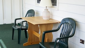 Une petite pièce avec une table en bois et deux chaises en plastique vertes. Sur la table, on trouve une lampe blanche, et il y a une fenêtre avec un rideau blanc.