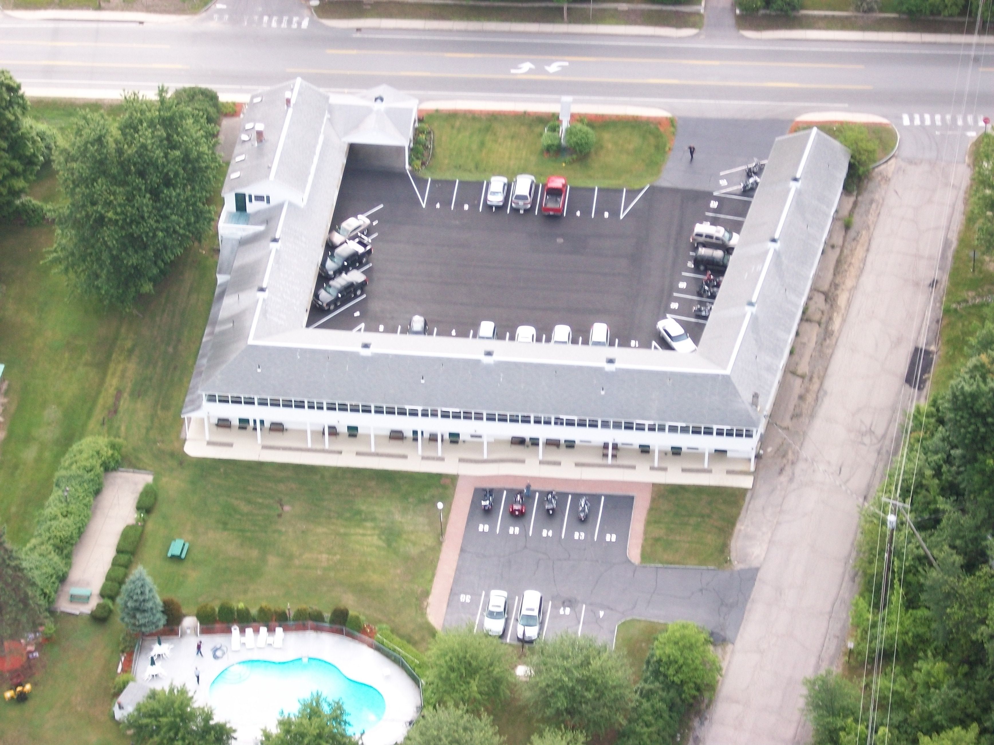 An aerial view of a building with a parking lot, swimming pool, and adjacent green spaces.