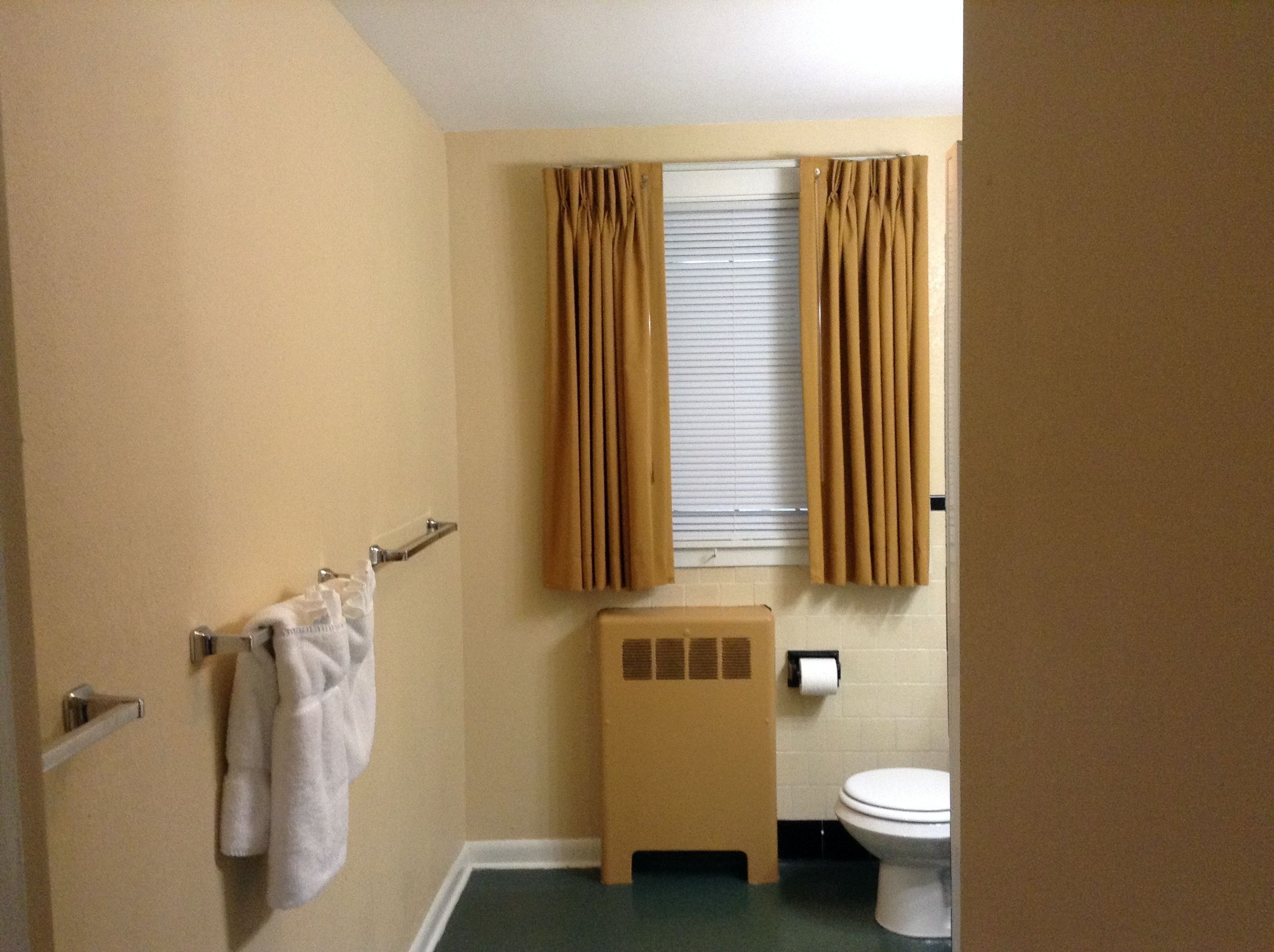 A bathroom with beige walls, a toilet, a towel rack with two towels, and a window with curtains.