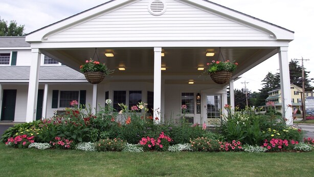 Un bâtiment blanc doté d’un porche abrité, de deux jardinières suspendues et d’un jardin fleuri à l’avant.