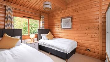 A wooden cabin bedroom with two beds, a nightstand, a lamp, and a window with curtains.