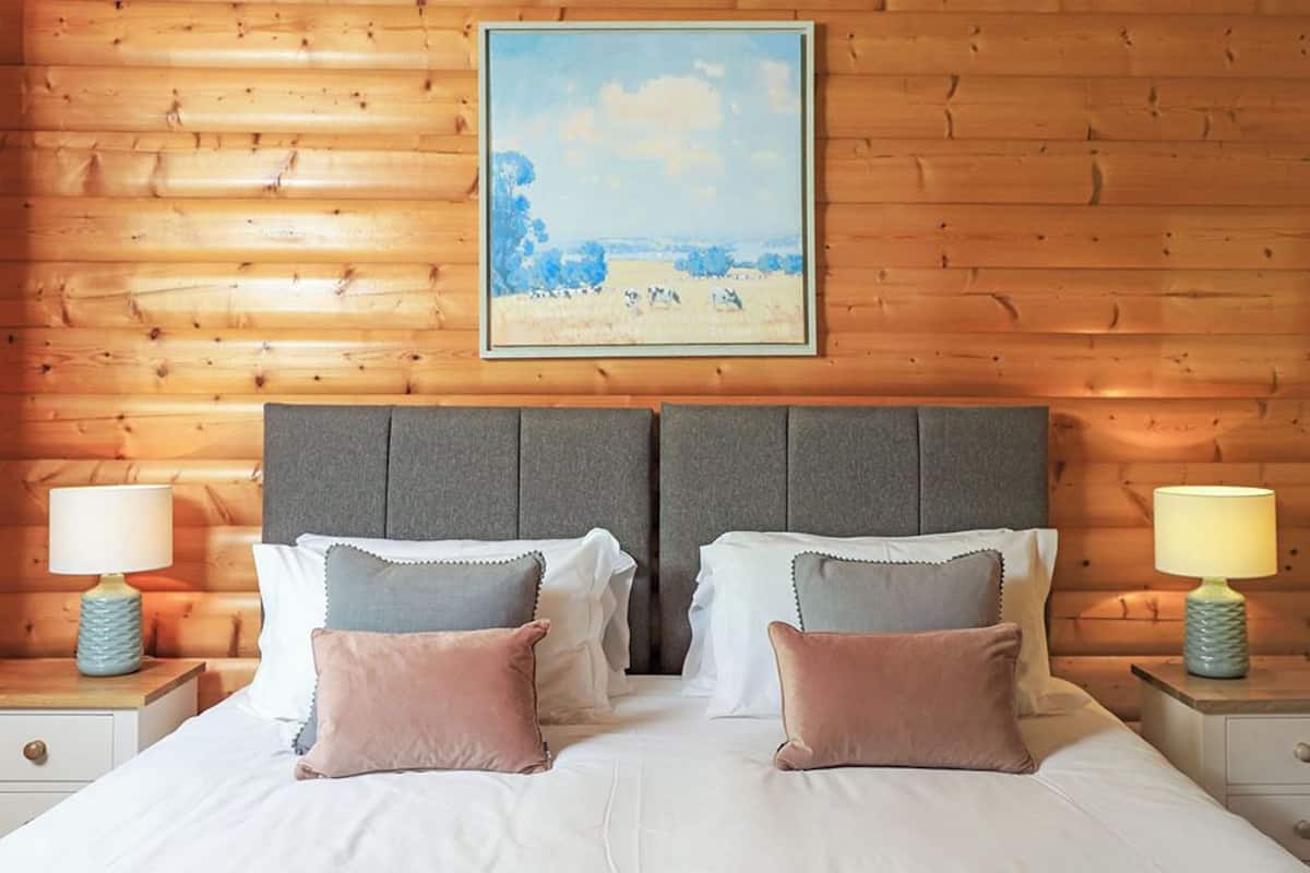 A double bed with grey headboard and white bedding, two bedside tables with lamps, and a painting of a landscape on the wall.