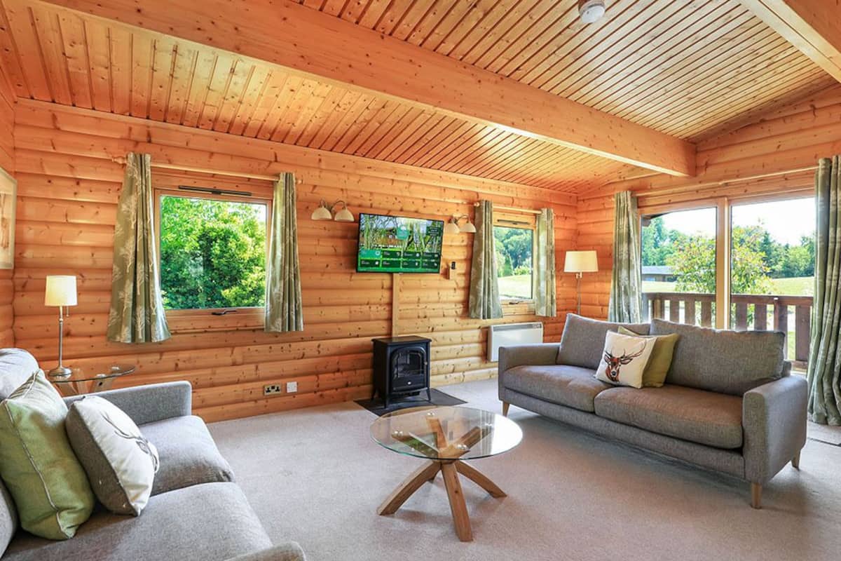 A cozy log cabin interior with a fireplace, a sofa, a television, and large windows offering a view of greenery.