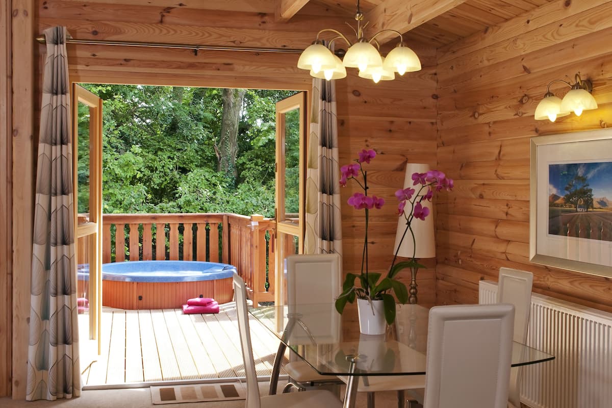 A wooden cabin with a glass table and chairs, a hot tub, and a view of a forested area.