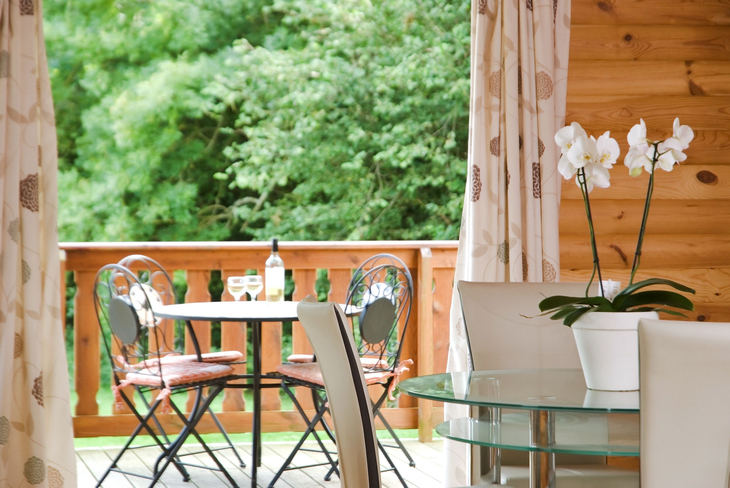 Eine Holzterrasse mit einem Glastisch und Stühlen, umgeben von Vorhängen und einem Blumentopf.