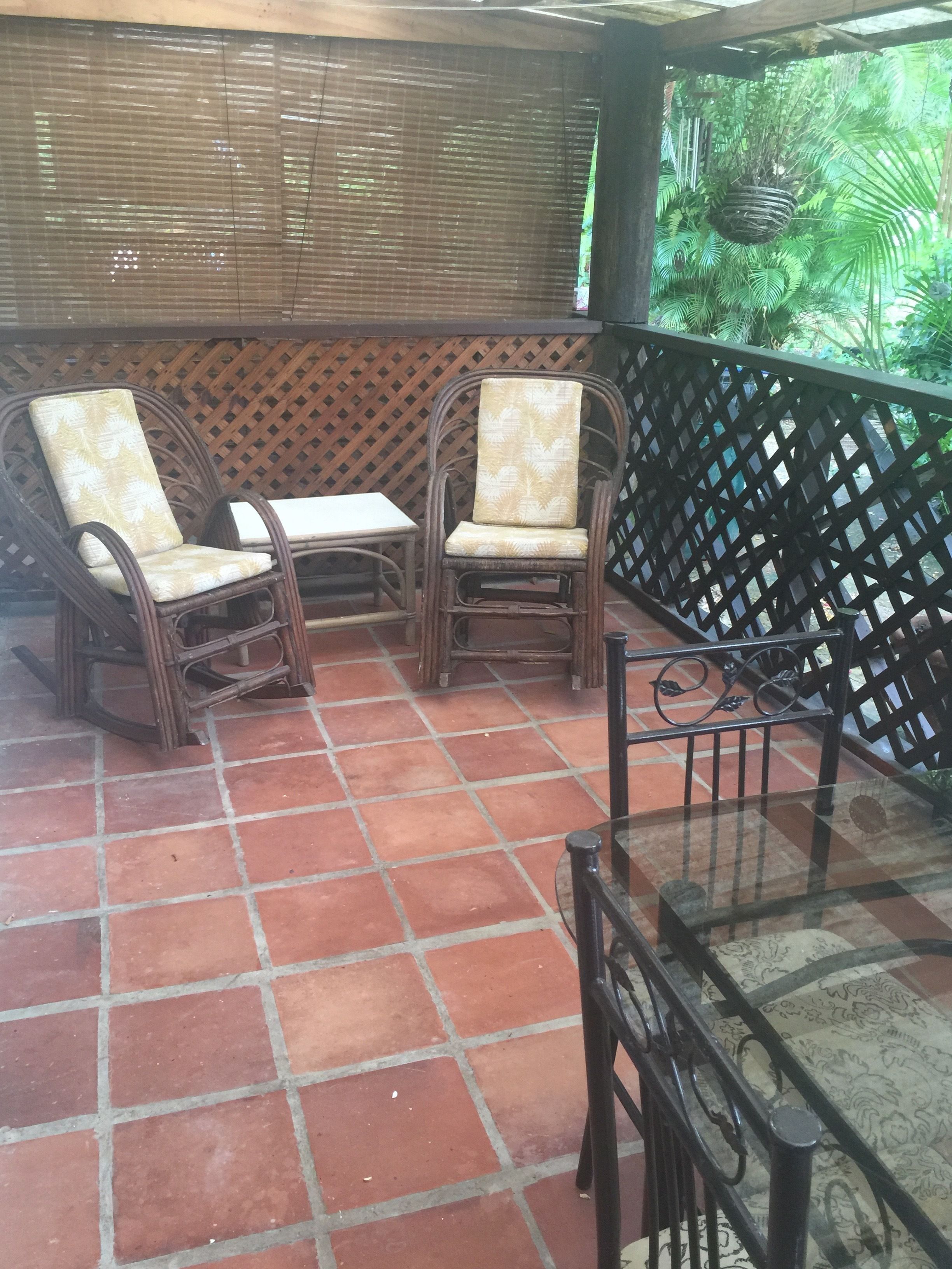 Un patio techado con mobiliario de mimbre, piso de baldosas y vista a la vegetación a través de una celosía.