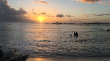 Escena de playa con barcos, agua tranquila y un atardecer.