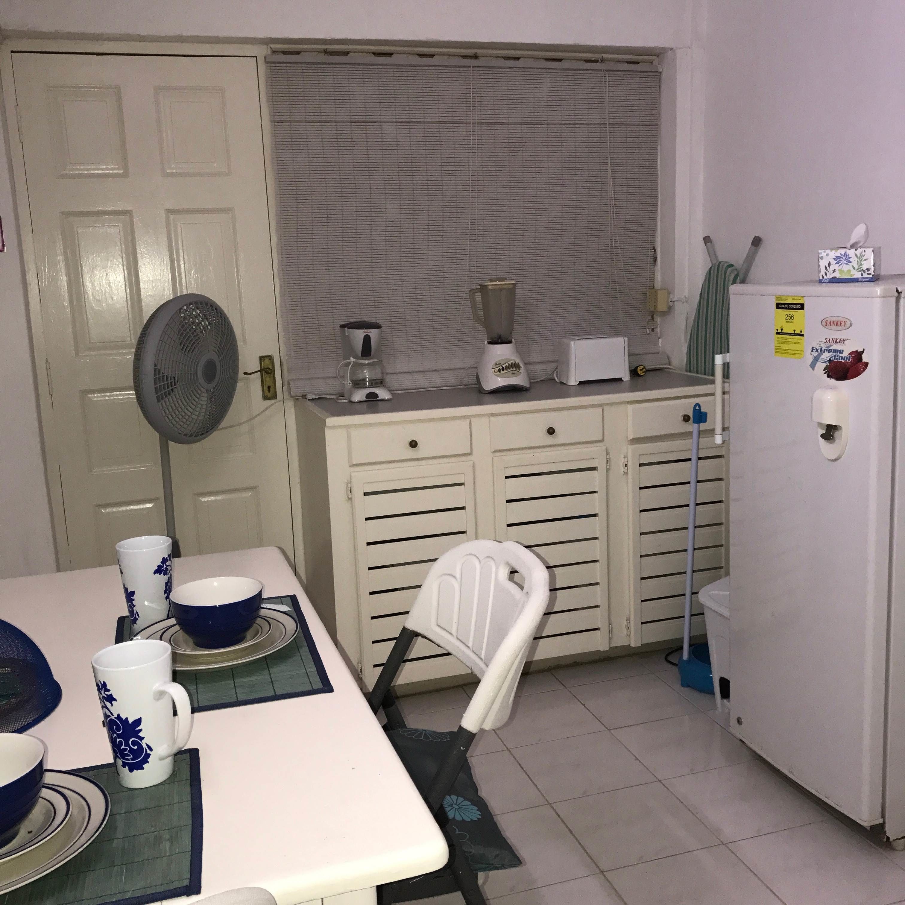 Una cucina compatta con frigorifero, macchina del caffè, frullatore e ventilatore. C'è un tavolo con una sedia e alcune tazze e ciotole.