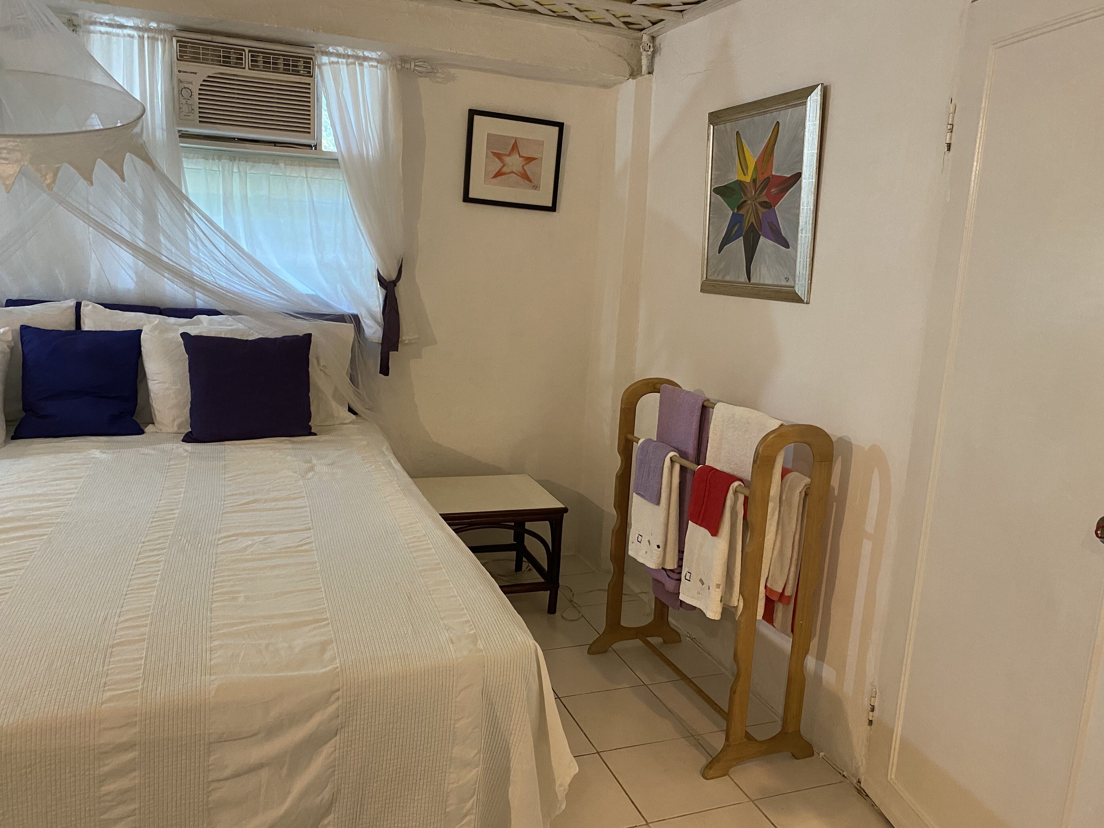 Un dormitorio con cama, mesita de noche, una silla, ventana con cortinas y una pared decorada con dos cuadros.
