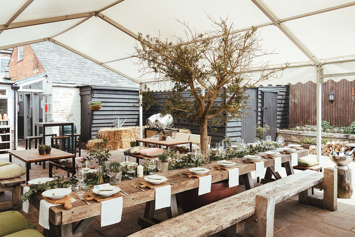 Un área de comedor al aire libre con una larga mesa de madera lista para una comida, rodeada de mobiliario rústico y un árbol.