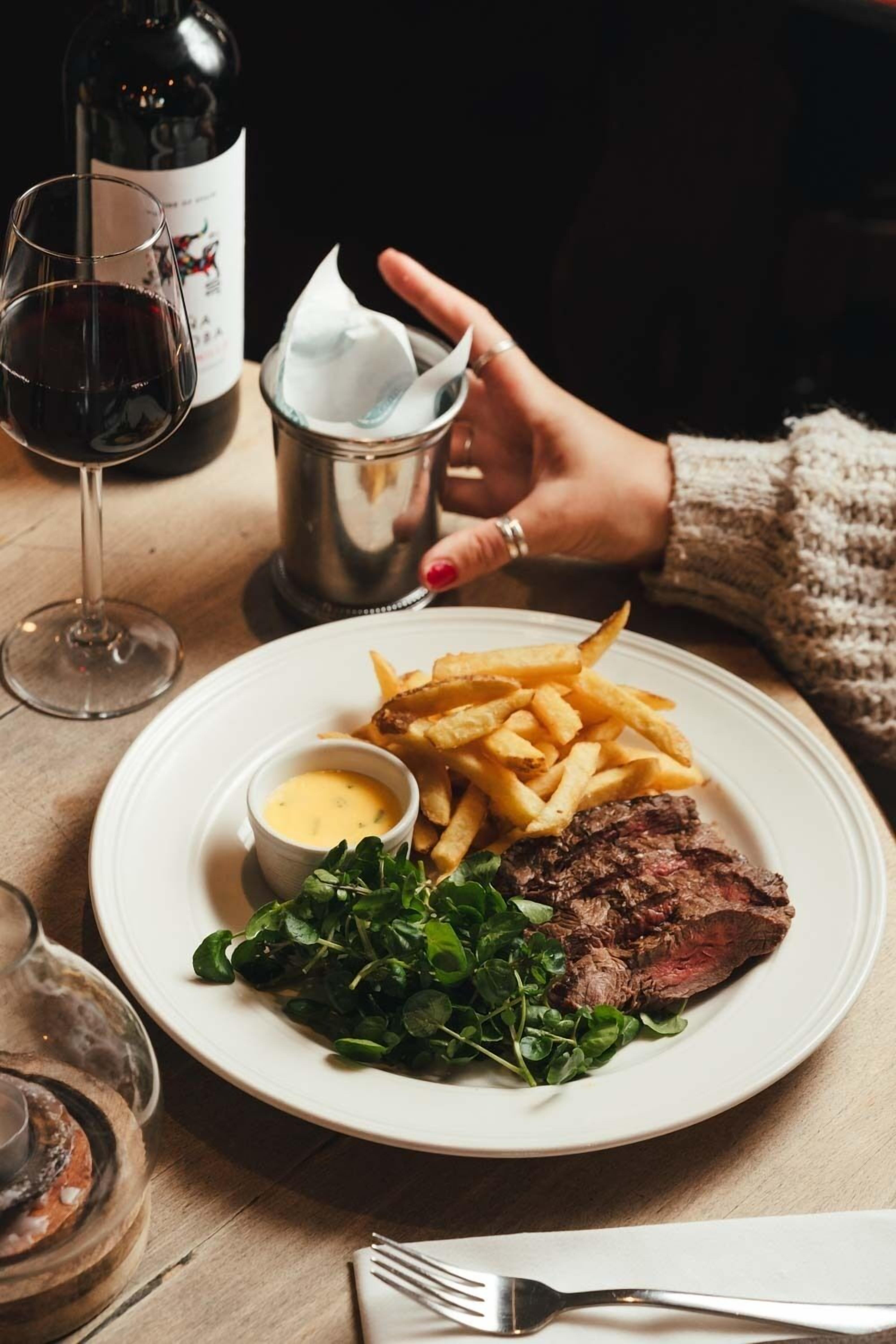 Een bord met steak, friet en groenten, een wijnglas en een fles wijn op een tafel.