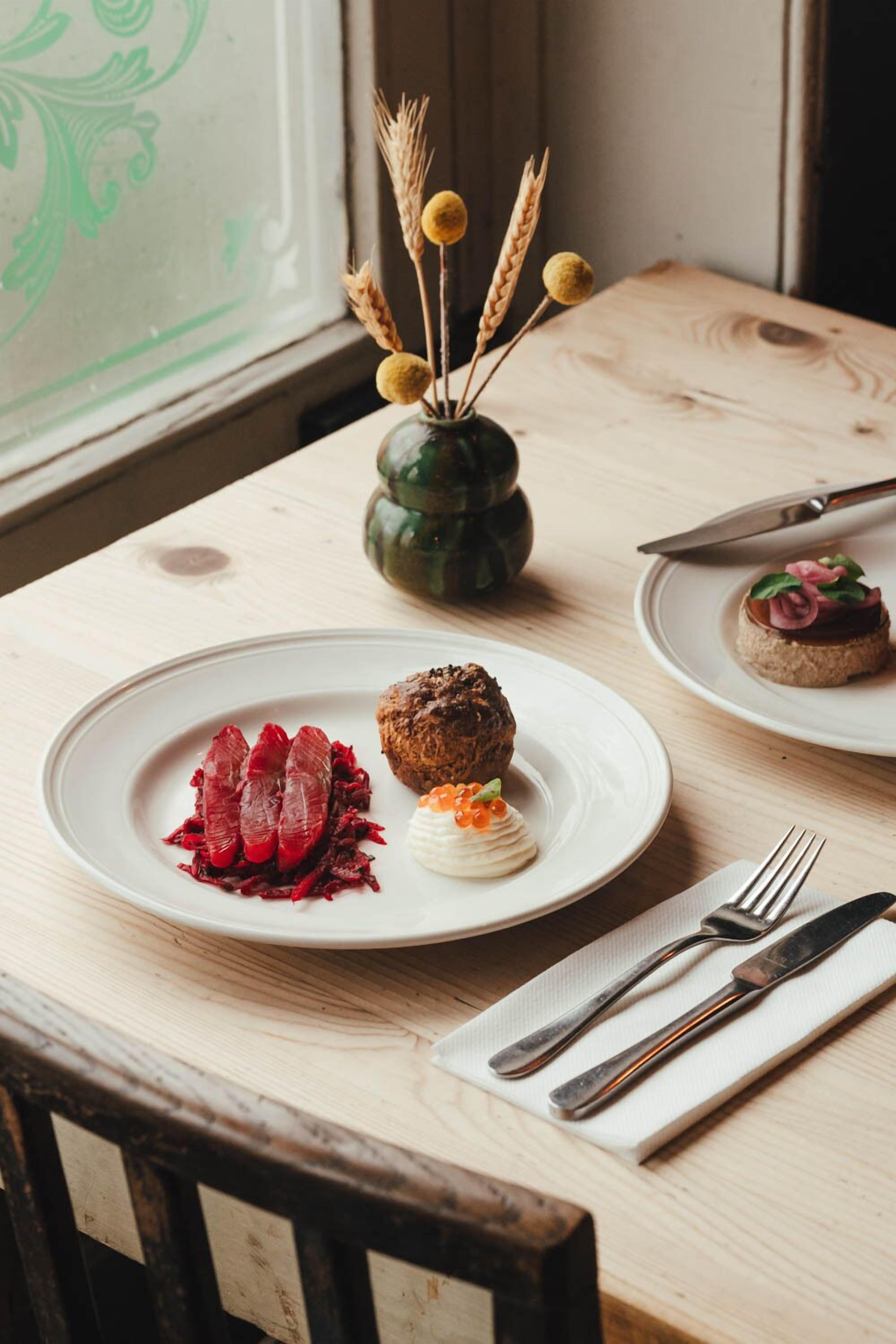A wooden table with plates of food, a vase with dried wheat stalks, and a window with a green design.