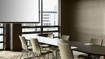 A conference room with a long table and six chairs, large windows with a city view, and a projector screen.